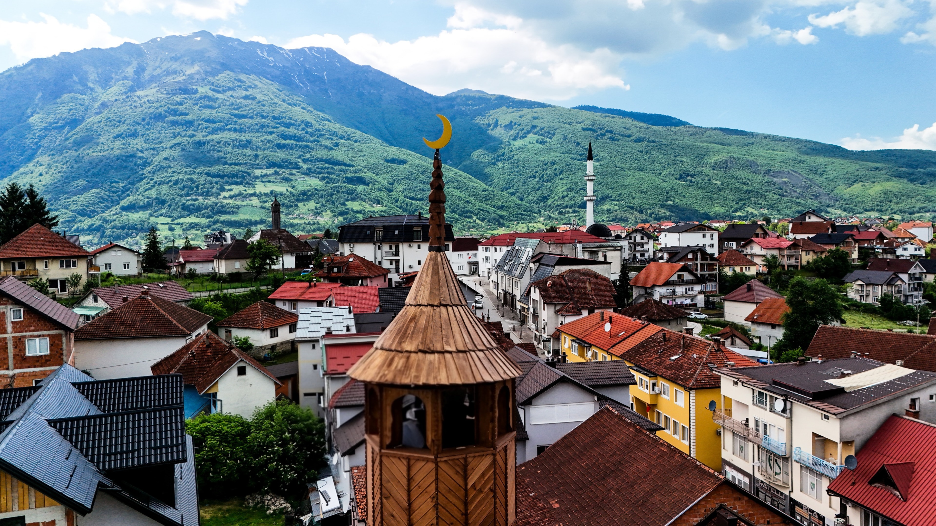 Drvena munara s polumjesecom iznad krovova Plava, sa zelenim planinama u pozadini